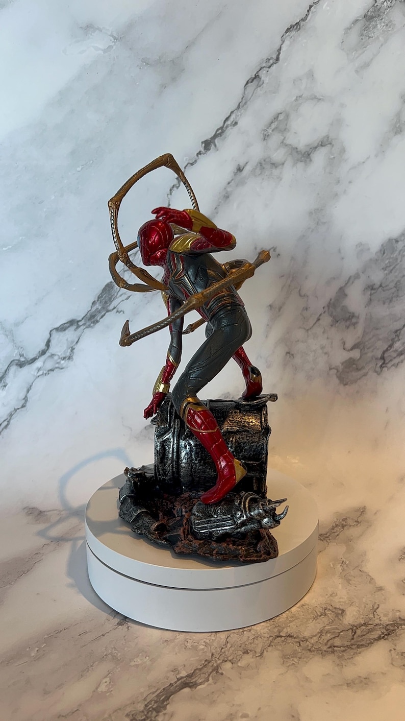 May include: A detailed figurine of a superhero in a dynamic pose, featuring a red and gold helmet and suit with black accents. The figure is mounted on a detailed base with metallic and dark brown tones, and includes multiple golden mechanical arms.