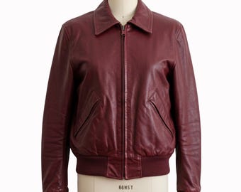 May include: A burgundy leather jacket with a collared neckline and a front zipper closure. The jacket features two front pockets and ribbed cuffs and hem. The jacket is displayed on a mannequin.