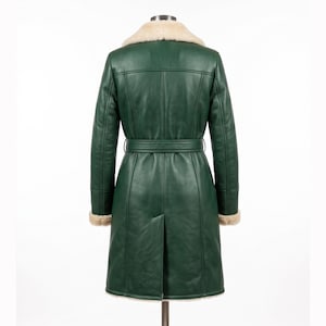 May include: A green leather coat with a cream-colored shearling collar and cuffs. The coat features a belt at the waist and a split back. The coat is displayed on a mannequin against a white background.