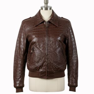 Puede incluir: Chaqueta bomber de cuero marrón con textura de cocodrilo. La chaqueta tiene cierre de cremallera, dos bolsillos delanteros y dobladillo y puños de canalé. La chaqueta se exhibe en un maniquí.