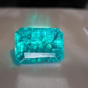 May include: A rectangular emerald-cut gemstone with a vibrant, translucent cyan-green color. The stone features a faceted surface that reflects light, creating internal patterns and a glowing effect across its crystalline structure.