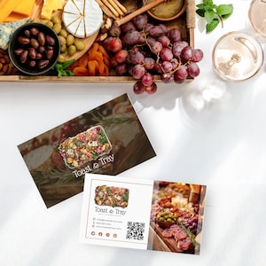 以下が含まれることがあります： チーズ、オリーブ、乾燥アプリコット、ブドウなど、さまざまな食材が載った木製トレイ。前面には2枚の名刺があり、1枚には「Toast & Tray」の文字が書かれています。もう1枚の名刺には、同じロゴと連絡先情報が記載されています。