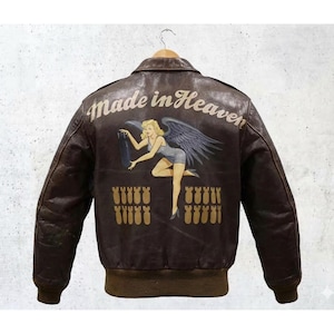 Puede incluir: Chaqueta bomber de cuero marrón con un diseño clásico. La parte trasera presenta un gráfico de chica pin-up con las palabras "Made in Heaven" encima. Debajo del gráfico hay filas de bombas amarillas. La chaqueta tiene cuello y puños acanalados marrones.