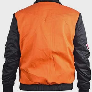 May include: Back view of a bomber jacket. The jacket features a bright orange body with black sleeves and a black collar. The jacket has a ribbed hem and cuffs. A small white patch with red text is on the left sleeve.
