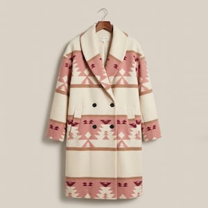 Puede incluir: Un abrigo cruzado crema con un estampado geométrico en tonos rosa y marrón. Tiene solapas anchas, dos filas de botones negros y dos bolsillos delanteros. El abrigo está colgado de una percha de madera.