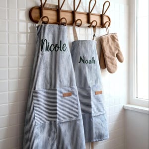Puede incluir: Dos delantales a rayas azules y blancas, bordados con los nombres "Nicole" y "Noah" en verde, colgados de un perchero de madera con un guante de horno beige. Los delantales tienen un bolsillo delantero y correas beiges.