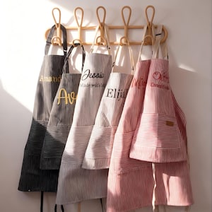 May include: Five personalized aprons with vertical stripes hang on a wooden rack. The aprons are in various colors: black, gray, and red. Each apron has a name embroidered on it in a contrasting color.