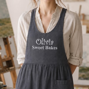 May include: A dark gray apron featuring the white embroidered text "Olivia Sweet Bakes" on the chest. The apron has a gathered waistline, a single front pocket, and wide shoulder straps, worn over a light beige long-sleeved button-down shirt.