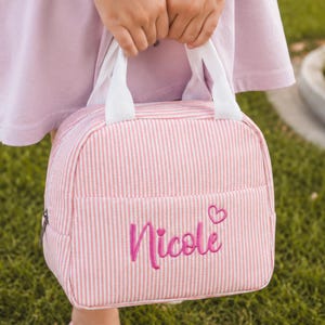 Puede incluir: Una bolsa de almuerzo a rayas rosas y blancas con asas de tela blanca y cierre de cremallera. La parte frontal de la bolsa presenta el nombre Nicole bordado en letra cursiva rosa, acompañado de un pequeño icono de corazón bordado en rosa.