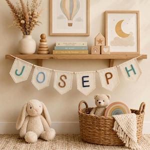 May include: A fabric banner featuring the name JOSEPH in felt letters, suspended below a wooden shelf. The shelf holds a vase with dried flowers, a wooden stacking toy, three books, and three wooden blocks. A plush bunny and a woven basket sit below.