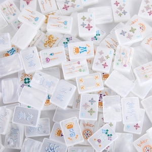 Puede incluir: Una colección de fichas de mahjong rectangulares blancas con varios diseños grabados en color, incluyendo números, patrones florales, coronas, la letra S, la letra W y la palabra Joker, todo con un acabado nacarado y suave.