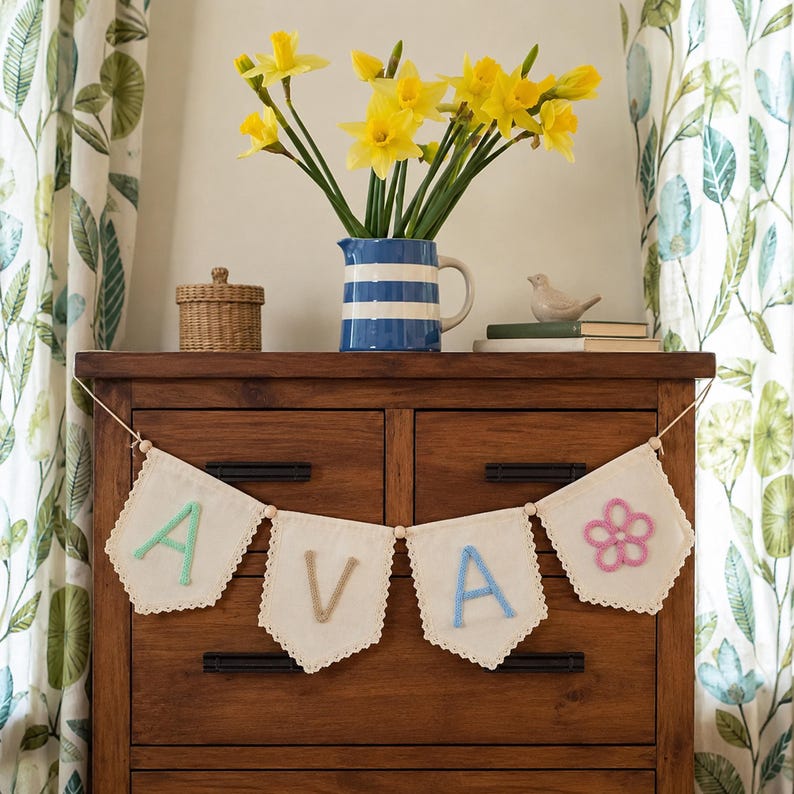 May include: A decorative fabric banner featuring four off-white pennants with scalloped edges, strung on a cord with wooden beads. The pennants display the letters A, V, A in pastel green, tan, and blue yarn, followed by a pink yarn flower.
