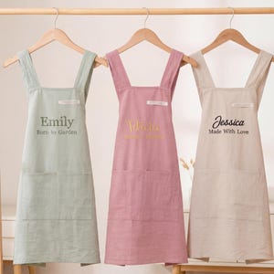 Puede incluir: Tres delantales personalizados colgados en un estante de madera. Los delantales son de color verde claro, rosa y beige. Cada delantal tiene un nombre diferente impreso, con textos como "Born to Garden" y "Made With Love".