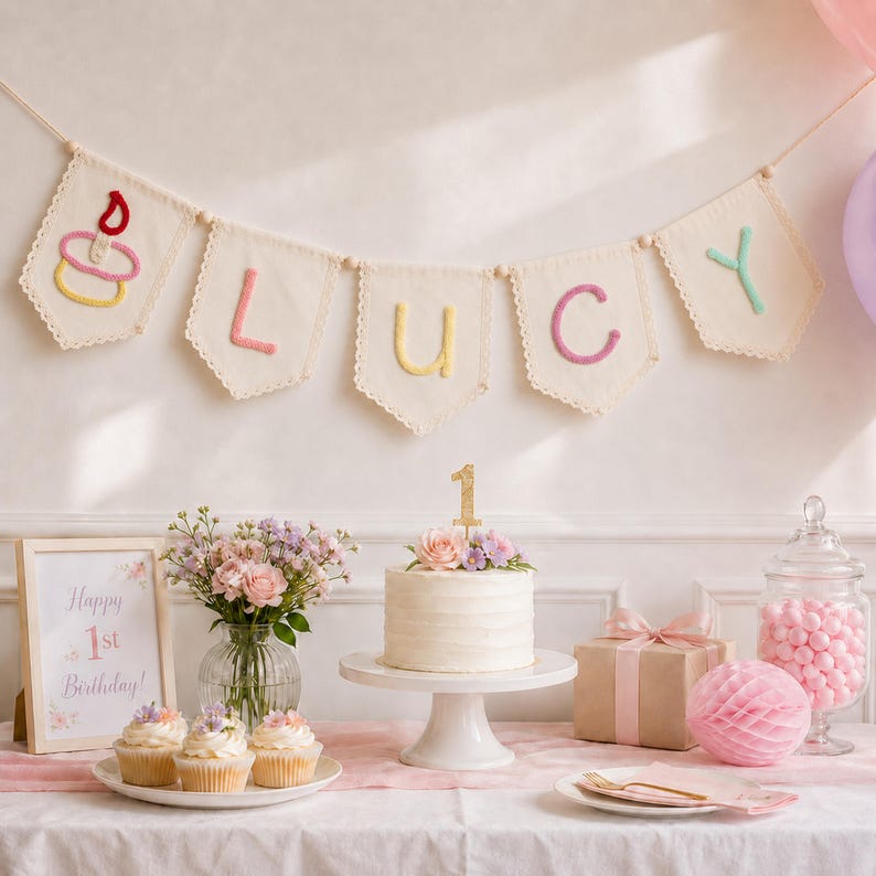 May include: A cream-colored fabric banner with scalloped edges features a stitched cupcake icon and the name LUCY in pastel yarn letters. Below, a white cake with a gold number 1 topper, cupcakes, a framed sign, and a gift box are displayed.