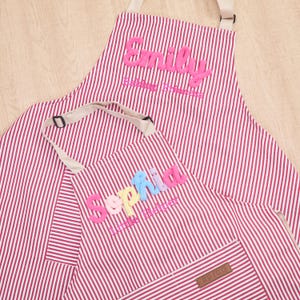 Puede incluir: Dos delantales con un patrón de rayas verticales rojas y blancas. El delantal más grande presenta el nombre Emily bordado en rosa y el texto Baking Princess. El más pequeño tiene el nombre Sophia en letras multicolores y Little Helper.