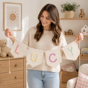 May include: A decorative fabric banner featuring four off-white pennants with scalloped edges, strung together with wooden beads. Each pennant displays a single raised letter in pastel colors, spelling out the name LUCY in pink, yellow, purple, and mint.