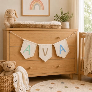 May include: A decorative fabric banner featuring three off-white pennants with scalloped edges, strung on a cord with wooden beads. The pennants display the letters A, V, and A in raised, soft-textured mint green, beige, and light blue yarn.