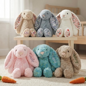 May include: Six soft toy bunny rabbits in shades of beige, blue, and pink. The bunnies have long ears with patterned fabric inside. Two toy carrots are placed in front of the bunnies. The bunnies are on a small wooden table and the floor.