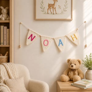 May include: A fabric bunting banner featuring four triangular pennants with scalloped edges, each displaying a single embroidered letter in pink, yellow, or blue, spelling the name NOAH, suspended by a white cord with wooden beads.