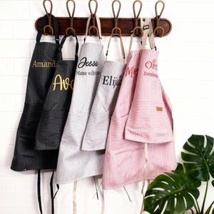 May include: A collection of personalized aprons hanging on a wooden rack. The aprons feature vertical stripes in black, gray, and pink, with embroidered names in various colors. Each apron has a pocket and ties.