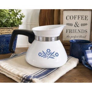 May include: A white ceramic teapot featuring a blue floral pattern on the front, a black handle, and a silver-toned metal band around the neck. The teapot has a wide, rounded base and a flared rim, designed for pouring hot beverages.