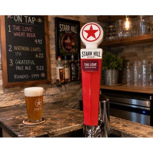 May include: A red and white Starr Hill Brewery beer tap handle featuring a circular top with a red star logo and a rectangular label reading The Love Wheat Beer, positioned next to a glass of beer with the same logo on a coaster.