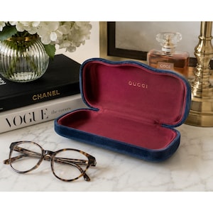 May include: An open, rectangular Gucci glasses case with a dark blue velvet exterior and a deep magenta interior lining. The brand name Gucci is printed in gold lettering on the inside. Beside the case are tortoise shell patterned eyeglasses.