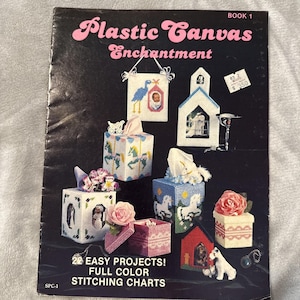Op de afbeelding: Een knutselinstructieboek getiteld Plastic Canvas Enchantment Book 1, met een zwarte omslag met afbeeldingen van diverse plastic canvas projecten, waaronder tissueboxhoezen, kleine doosjes en wandhangers met bloem- en diermotieven.