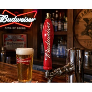 May include: A red Budweiser beer tap handle with white lettering, positioned next to a clear glass of golden beer featuring a white foam head and the Budweiser logo, set on a wooden bar top with a neon Budweiser sign in the background.