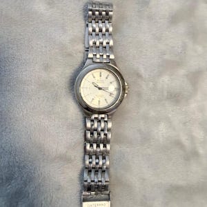 Op de afbeelding: Een zilverkleurig analoog polshorloge met een ronde wijzerplaat, een witte achtergrond en een metalen schakelarmband. Op de wijzerplaat staan de teksten GUESS en WATERPRO, met zilveren uurmarkeringen en wijzers. De horlogekast is van gepolijst metaal.