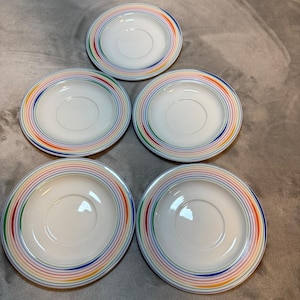 Puede incluir: Cinco platillos blancos con rayas concéntricas de colores del arcoíris alrededor del borde. Los platillos tienen una hendidura central para una taza. Los colores incluyen rojo, naranja, amarillo, verde, azul y morado. Los platillos están dispuestos sobre una superficie de color neutro.