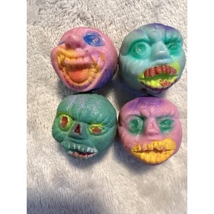 May include: Four small, round, sculpted heads with exaggerated facial expressions. One is pink and orange, one is teal and purple, one is green with red-rimmed eyes, and one is pink and purple with a yellow mouth. Each has distinct, textured features.