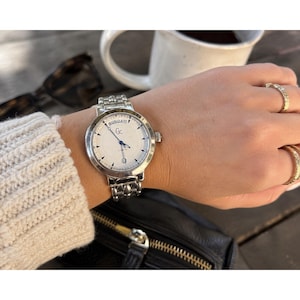 Peut inclure: Une montre-bracelet analogique argentée avec un cadran rond blanc, arborant le logo Gc, un affichage du jour de la semaine en haut et un guichet de date en bas. La montre possède un bracelet à maillons en métal poli et des aiguilles fines.