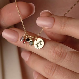 May include: A gold-colored necklace with a delicate chain and multiple charms. The charms include small gemstones in various colors and a round pendant engraved with the name "gigi". The necklace is held by a person.