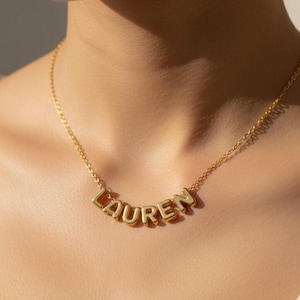 Könnte beinhalten: Eine goldfarbene Halskette mit dem Namen "LAUREN" in Großbuchstaben. Die Kette hängt an einer feinen Kette. Die Buchstaben sind gleichmäßig angeordnet und leicht gebogen. Die Kette wird auf der Haut einer Person getragen.