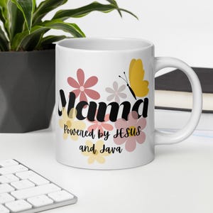 Puede incluir: Taza de cerámica blanca con la palabra "Mama" en escritura negra, decorada con flores rosas y amarillas y una mariposa amarilla. El texto "Powered by JESUS and Java" también está en la taza.