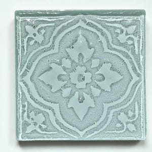 May include: Square, light blue glass tile with an etched floral design. The design is symmetrical, featuring a central flower and corner accents. The tile has a slightly textured surface and a clear border.