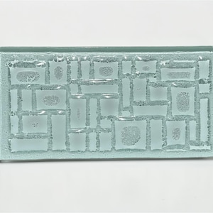 May include: A rectangular, light blue glass decorative piece with a textured surface. The surface features a pattern of various-sized rectangles and squares. The glass has a frosted appearance, and the edges are smooth.