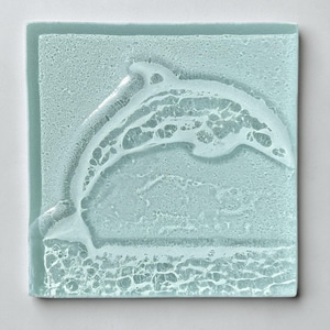 May include: A square, light blue glass coaster with a raised dolphin design. The dolphin is leaping out of the water, with textured details representing waves and bubbles. The coaster has a textured surface.