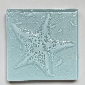 May include: A square glass tile in a pale, translucent blue color featuring a raised, textured white starfish design in the center. The surface of the glass has a smooth, glossy finish with a subtle, irregular pattern that mimics the appearance of water.