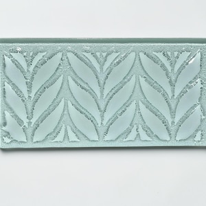 May include: A rectangular, light blue glass tile with a leaf pattern. The leaves are etched into the glass, creating a frosted effect. The tile is likely for use in a kitchen or bathroom as a decorative element.