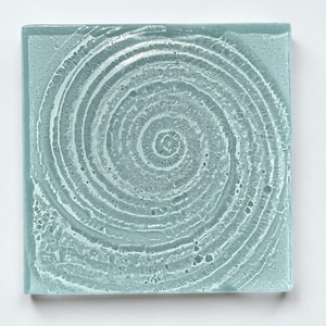May include: Square, light blue glass coaster with a white spiral design. The coaster has a textured surface and a slightly irregular edge. The spiral pattern starts from the center and expands outwards.