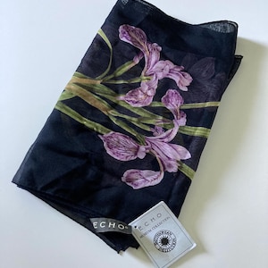 Puede incluir: Bufanda negra con un diseño floral de iris moradas y tallos verdes. La bufanda tiene una etiqueta que dice "ECHO MUSEUM COLLECTION SMITHSONIAN INSTITUTION".