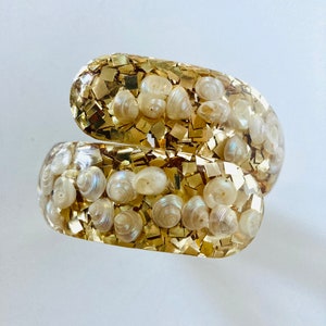 May include: A gold-colored resin bracelet with a swirl design. The bracelet is filled with small seashells and gold-colored glitter. The bracelet is a unique piece of jewelry.