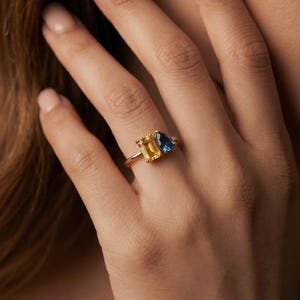 May include: A gold ring featuring two distinct gemstones: a rectangular emerald-cut yellow stone and a smaller pear-shaped blue stone set side-by-side on a thin gold band. The text SparkleCraftUSA is visible in the bottom right corner.