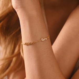 Op de afbeelding: Een delicate gouden armband met de namen "Winter" en "Roman" in cursief schrift. De armband wordt om de pols van een persoon gedragen, met de namen verbonden door een fijne ketting. Een gepersonaliseerd sieraad.