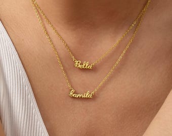 May include: A layered gold-colored necklace with two chains. The top chain features the name "Bella" in cursive script, and the bottom chain displays the name "Kamila". The necklaces are worn against a white top.