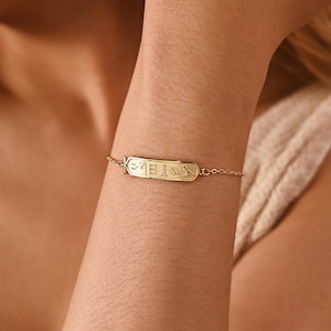 Könnte beinhalten: Ein goldfarbenes Armband mit einem rechteckigen Anhänger mit eingravierten Symbolen. Das Armband hat eine feine Kette. Der Anhänger befindet sich mittig am Handgelenk. Das Design ist minimalistisch und elegant.