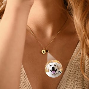 May include: A gold-colored necklace with a heart-shaped pendant and a small gemstone. The heart pendant has the letter "B" on it. A circular image of a dog is displayed below the necklace. The text "SparkleCraftUSA" is visible.