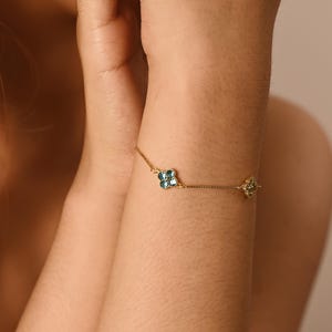 Könnte beinhalten: Ein filigranes goldenes Armband mit zwei floralen Motiven. Eines ist ein vierblättriges Kleeblatt-Design mit teal-farbenen Steinen, das andere ein kleineres, ähnliches Design mit grünen Steinen. Das Armband wird am Handgelenk getragen.